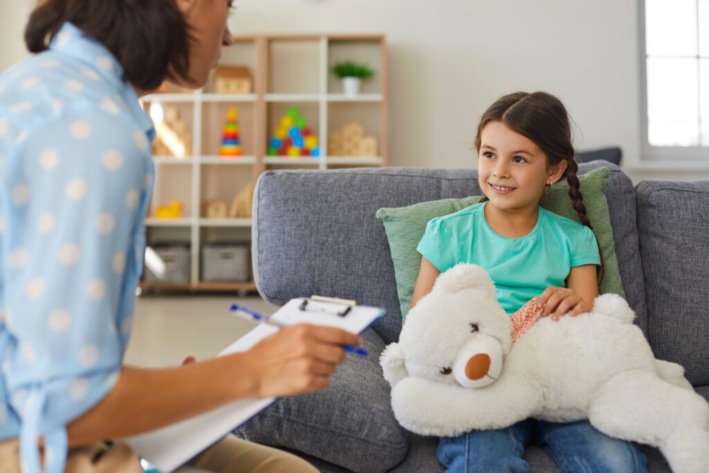 Supportive psychologist with clipboard talking to little girl. Therapist and happy child communicating, analyzing behavior and solving problems during therapy session in cozy modern office